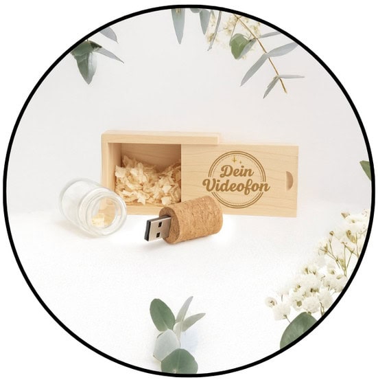 Eine Holzkiste mit der Aufschrift Dein Videofon mit Holzspänen, ein USB-Laufwerk aus Kork und ein kleines Glasgefäß, umgeben von weißen Blumen und grünen Blättern.
