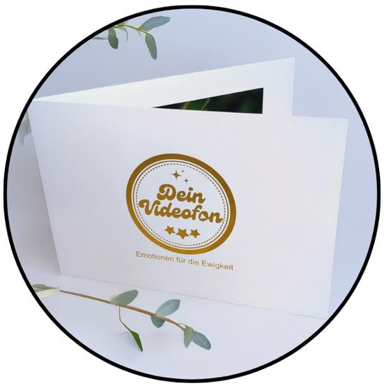 Eine weiße Grußkarte mit goldenem Text "Dein Videofan" und "Emotionen für die Ewigkeit", teilweise geöffnet und mit einem Zweig Grünzeug in der Nähe.